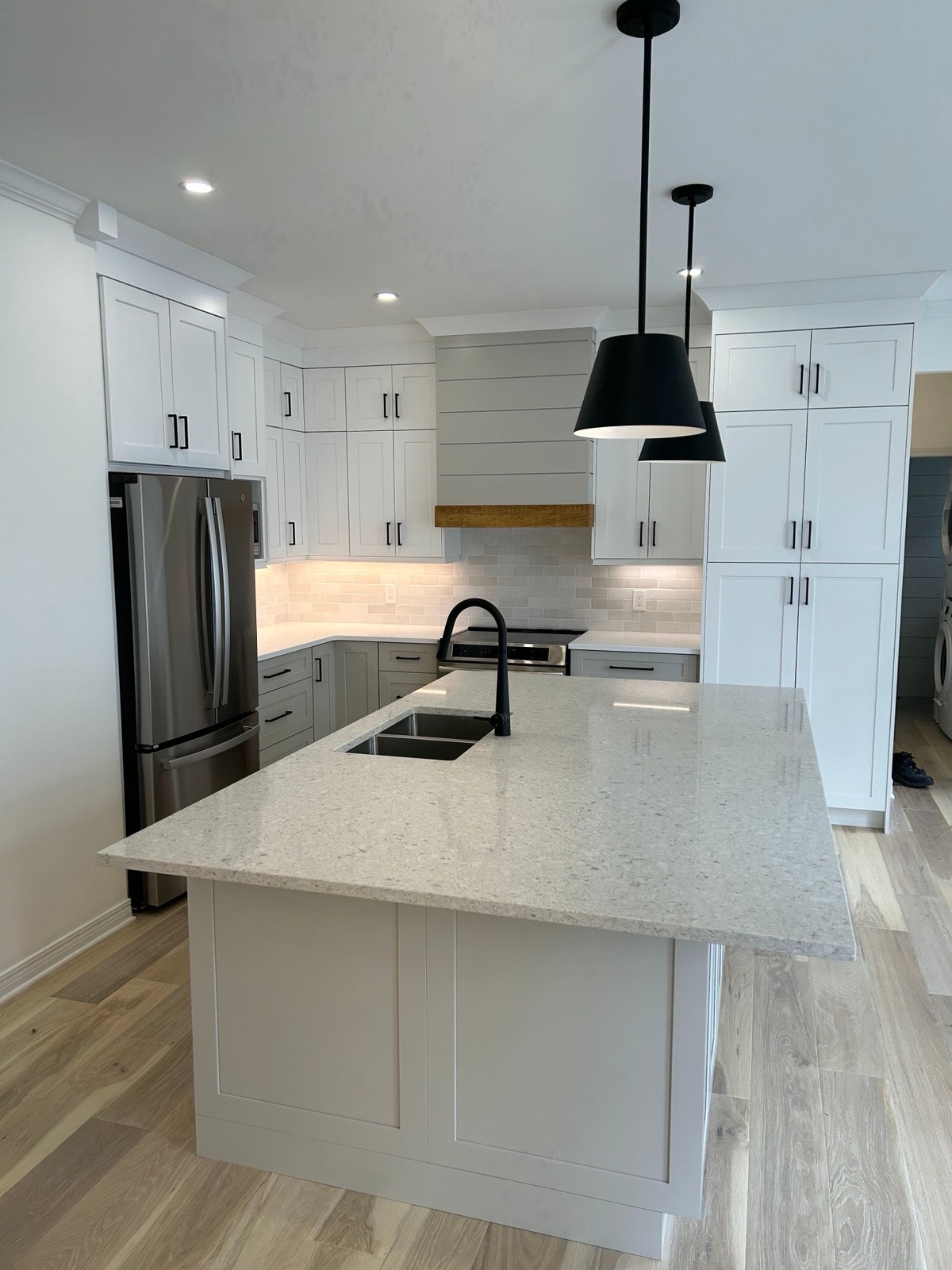 White shaker kitchen with large quartz island, matte black sink faucet, wood-trimmed range hood, black hanging lights and light hardwood flooring